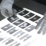 Mit Handschuhen behandelte Hände untersuchen fotografische Filmnegative auf einem Leuchttisch. (KI generierte Beschreibung)