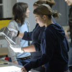 Mehrere Personen stehen um einen Tisch herum und begutachten fotografische Abzüge; eine Person trägt weiße Handschuhe, während sie ein Foto hält. (KI generierte Beschreibung)