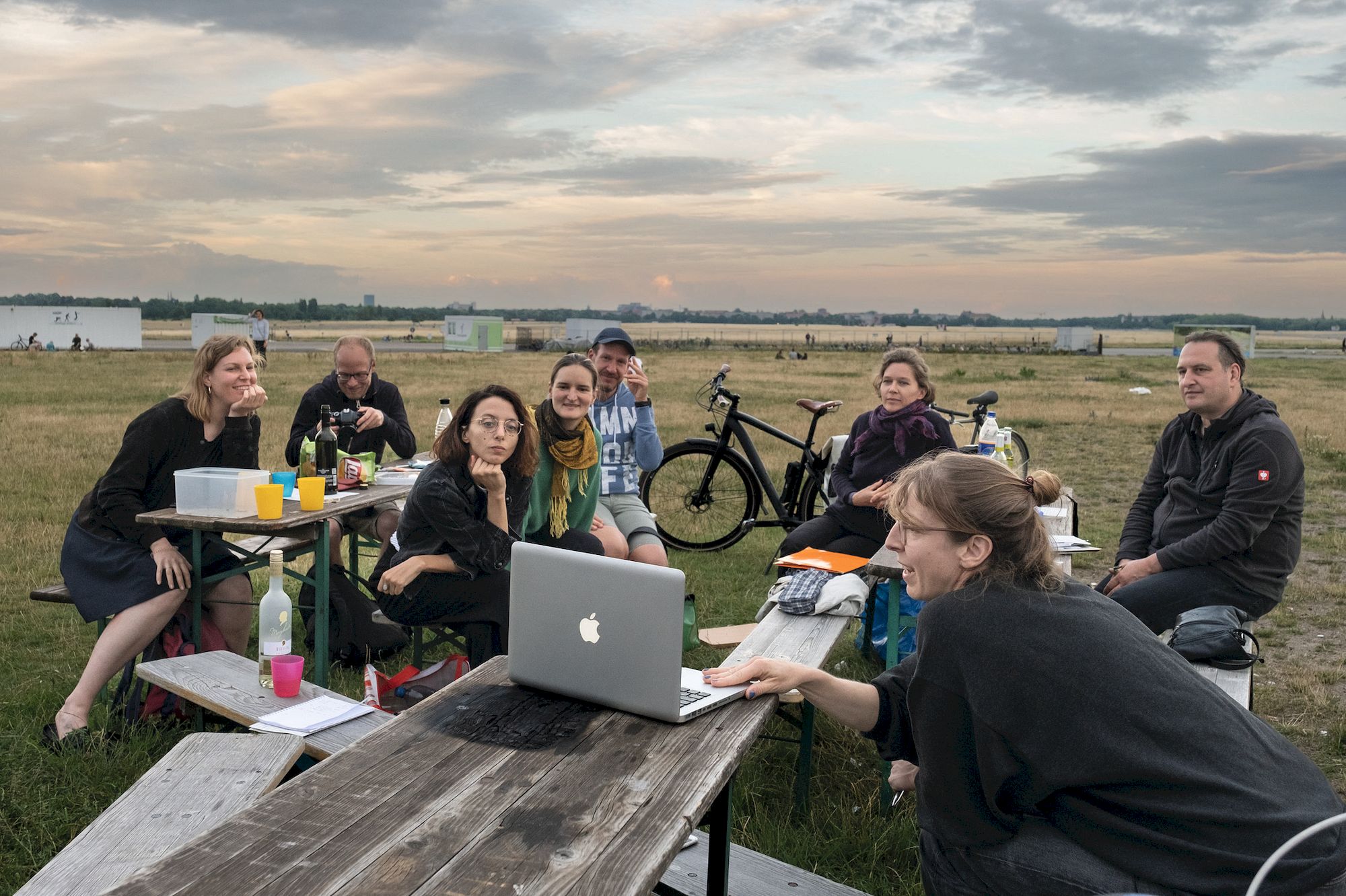 Eine Gruppe von Menschen sitzt um einen Picknicktisch und schaut gemeinsam Bilder auf einem Laptop an.