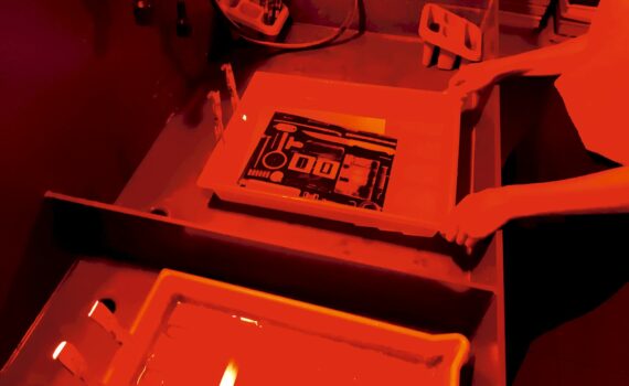 Eine Person arbeitet bei rotem licht in einer Dunkelkammer. In einer Schale mit Flüssigkeit liegt ein Schwarzweiß-Bild.