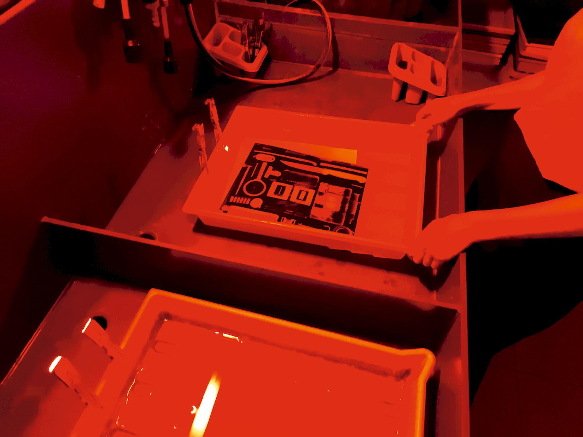 Eine Person arbeitet bei rotem licht in einer Dunkelkammer. In einer Schale mit Flüssigkeit liegt ein Schwarzweiß-Bild.