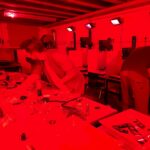 Eine Gruppe von Menschen, die in einer großen Dunkelkammer arbeiten. Alles ist in rotes Licht getaucht. Im Hintergrund sind sechs Arbeitsplätze mit Vergrößerern zu erkennen.