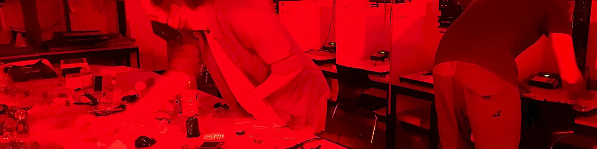 Eine Gruppe von Menschen, die in einer großen Dunkelkammer arbeiten. Alles ist in rotes Licht getaucht. Im Hintergrund sind sechs Arbeitsplätze mit Vergrößerern zu erkennen.