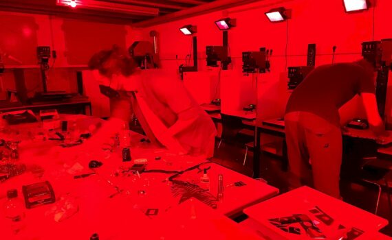 Eine Gruppe von Menschen, die in einer großen Dunkelkammer arbeiten. Alles ist in rotes Licht getaucht. Im Hintergrund sind sechs Arbeitsplätze mit Vergrößerern zu erkennen.