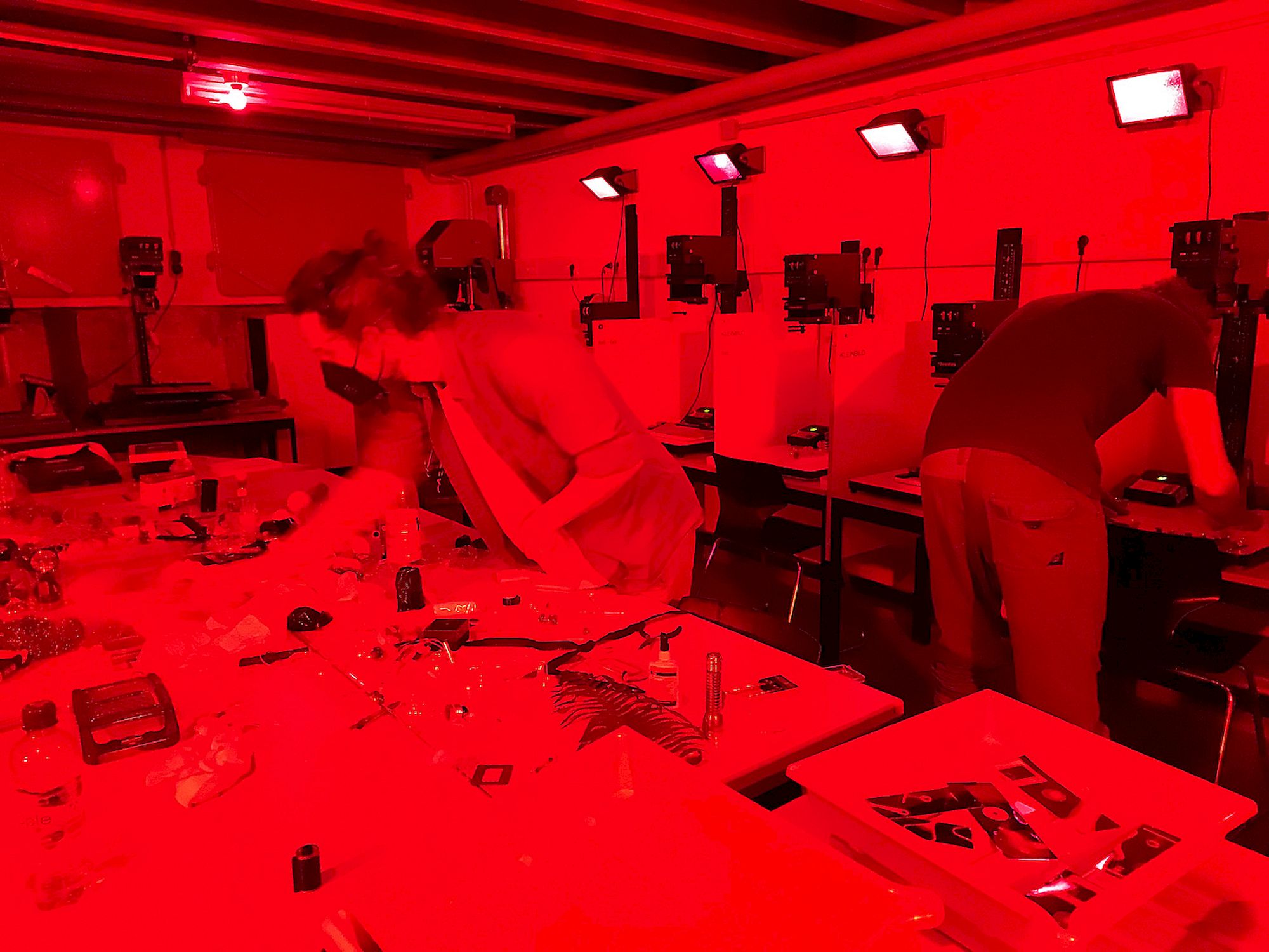 Eine Gruppe von Menschen, die in einer großen Dunkelkammer arbeiten. Alles ist in rotes Licht getaucht. Im Hintergrund sind sechs Arbeitsplätze mit Vergrößerern zu erkennen.