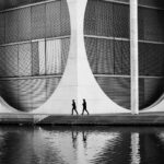 Ein Schwarz-Weiß-Foto von zwei Menschen, die vor einem Gebäude mit runden Fenstern gehen.