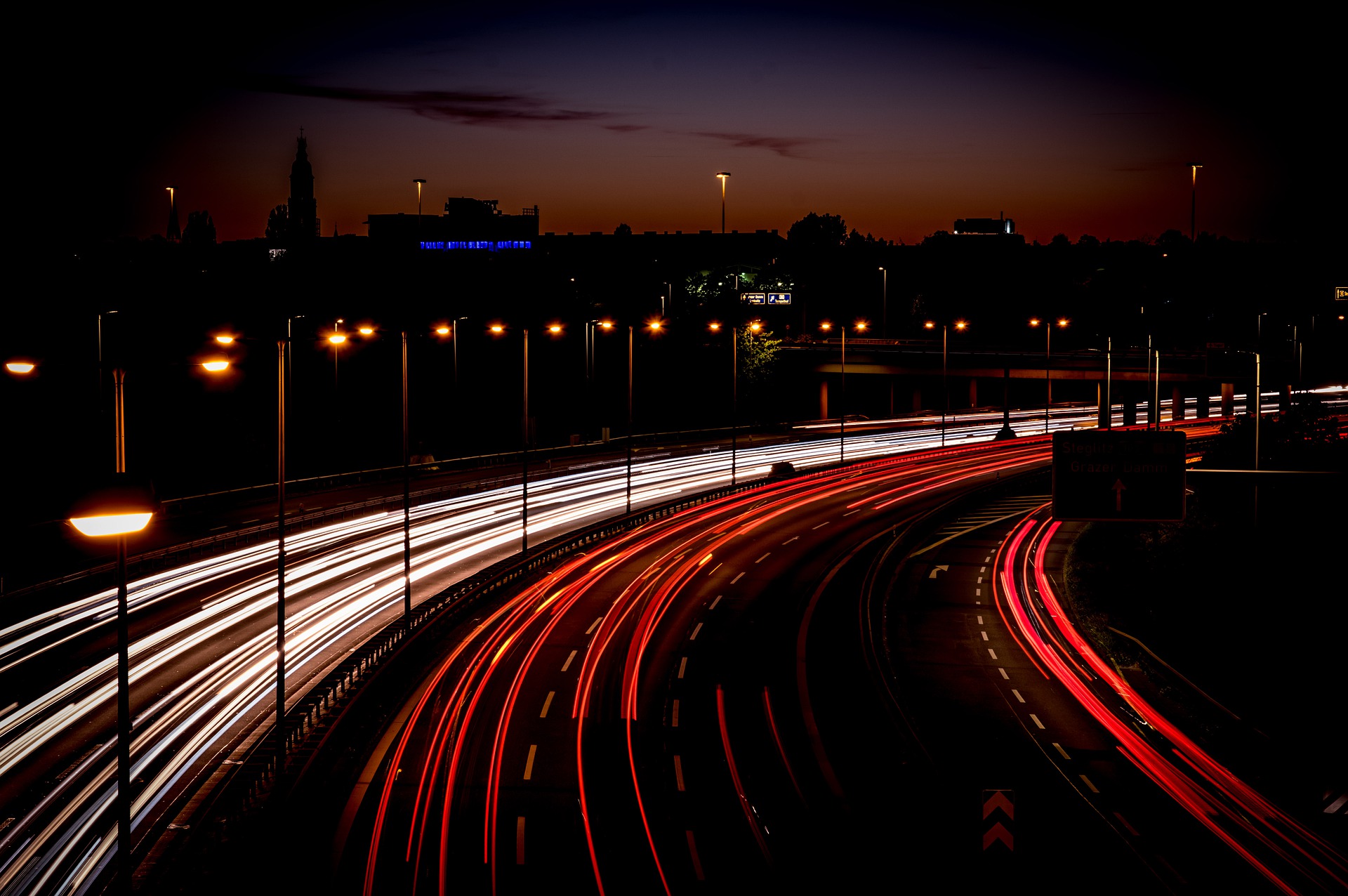Eine Langzeitbelichtungsaufnahme einer Stadt bei Nacht mit Lichtspuren von Autos in einer Kurve..