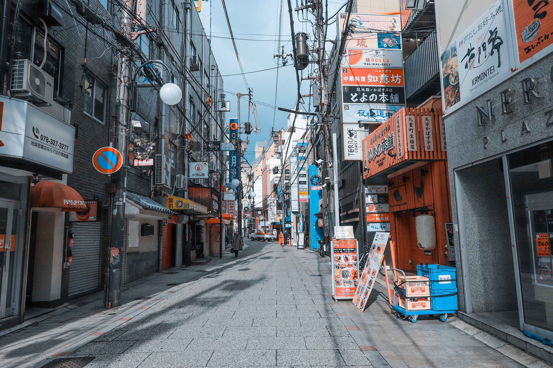 Eine menschenleere Straßenflucht in der japanischen Stadt Kobe. Oberleitungen werfen Schatten auf den sonnenbeschienen Boden. An den Häusern sind viele Werbetafeln angebracht.