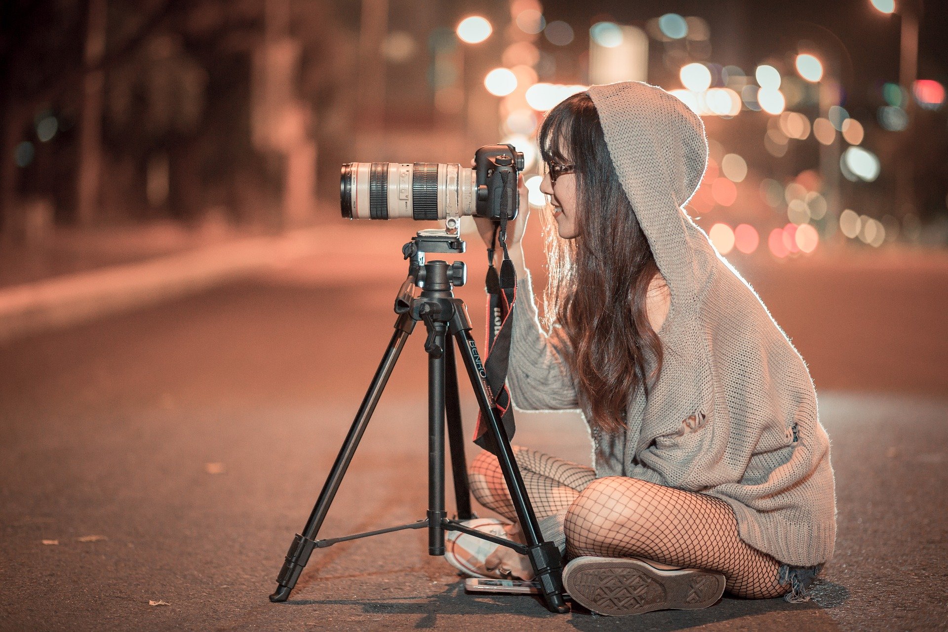 Eine junge Frau mit Kamera und Stativ sitzt nachts auf dem Boden und fotografiert mit einem Teleobjektiv.