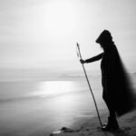 Ein Schwarz-Weiß-Foto einer Frau mit einem Stock am Strand. Der weite Mantel mit Kapuze weht im Wind. Im Hintergrund sind Berge zu sehen.