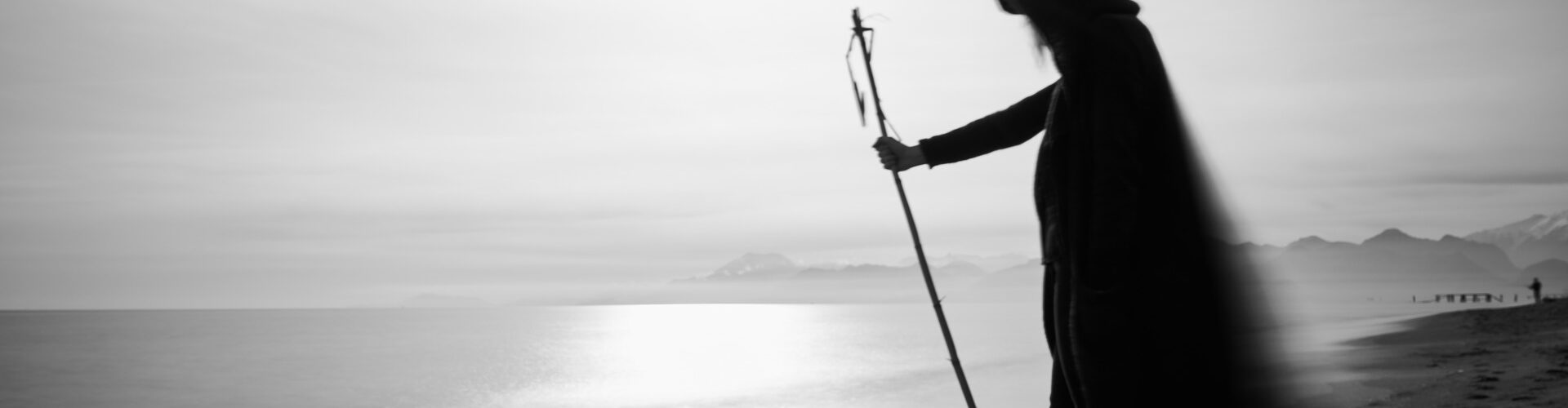 Ein Schwarz-Weiß-Foto einer Frau mit einem Stock am Strand. Der weite Mantel mit Kapuze weht im Wind. Im Hintergrund sind Berge zu sehen.