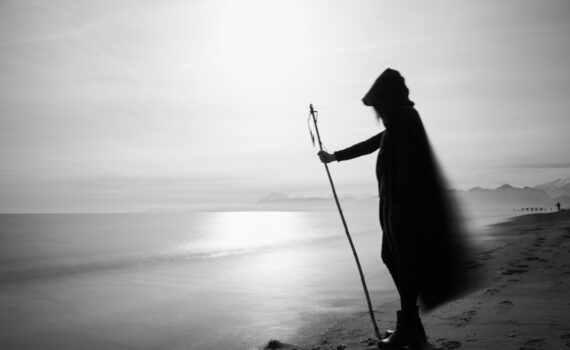 Ein Schwarz-Weiß-Foto einer Frau mit einem Stock am Strand. Der weite Mantel mit Kapuze weht im Wind. Im Hintergrund sind Berge zu sehen.
