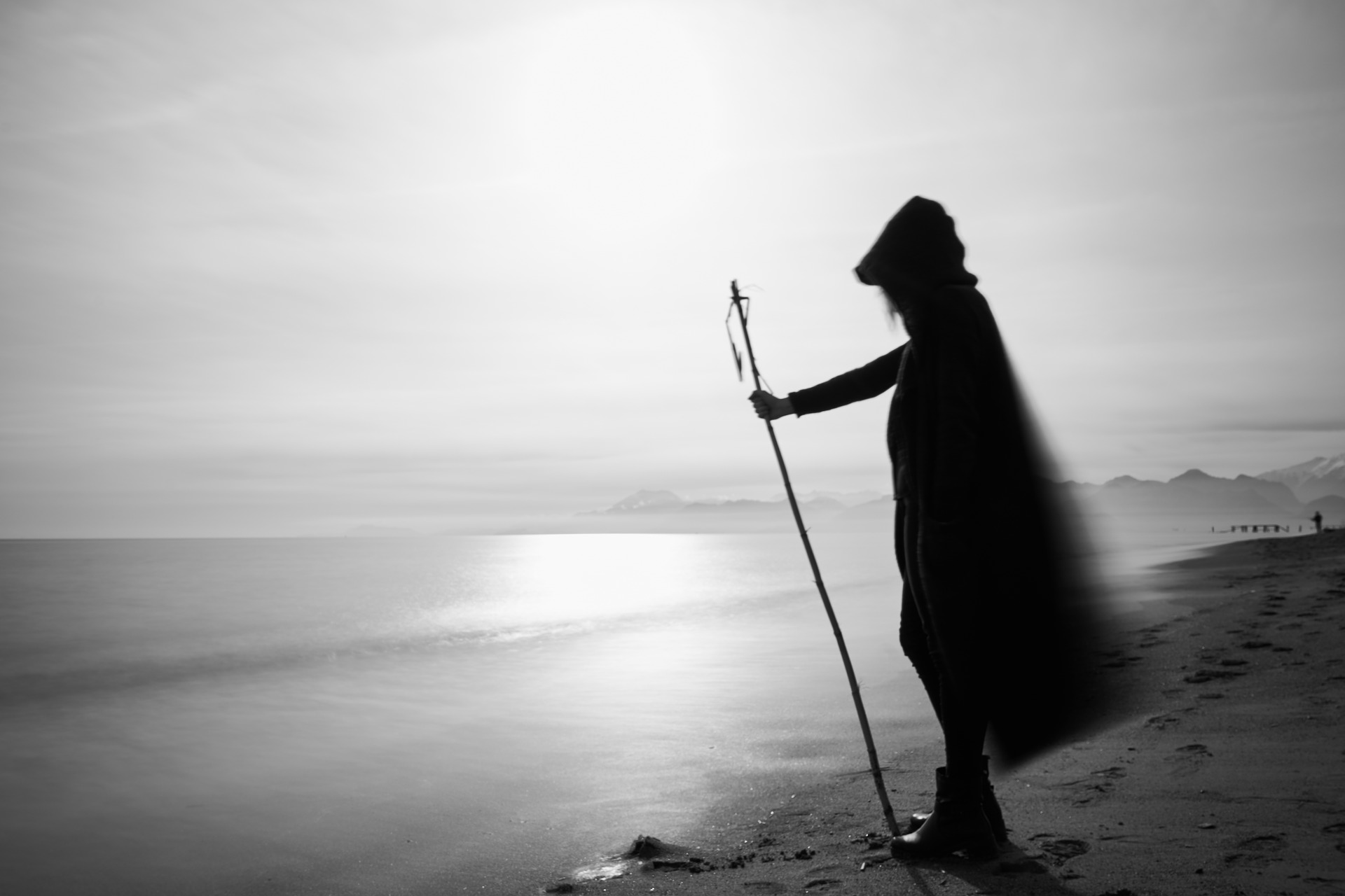 Ein Schwarz-Weiß-Foto einer Frau mit einem Stock am Strand. Der weite Mantel mit Kapuze weht im Wind. Im Hintergrund sind Berge zu sehen.