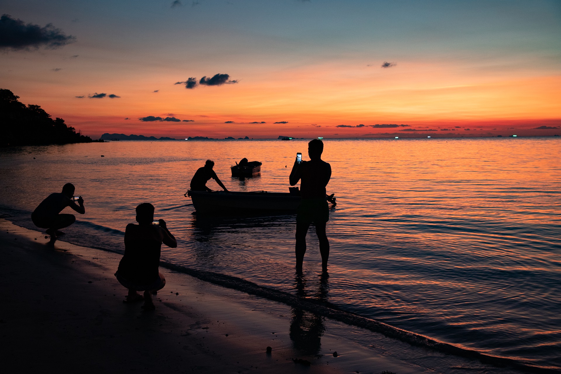 Eine Gruppe von Menschen steht bei Sonnenuntergang am Strand und fotografiert mit dem Handy einen Fischer mit seinem Boot.