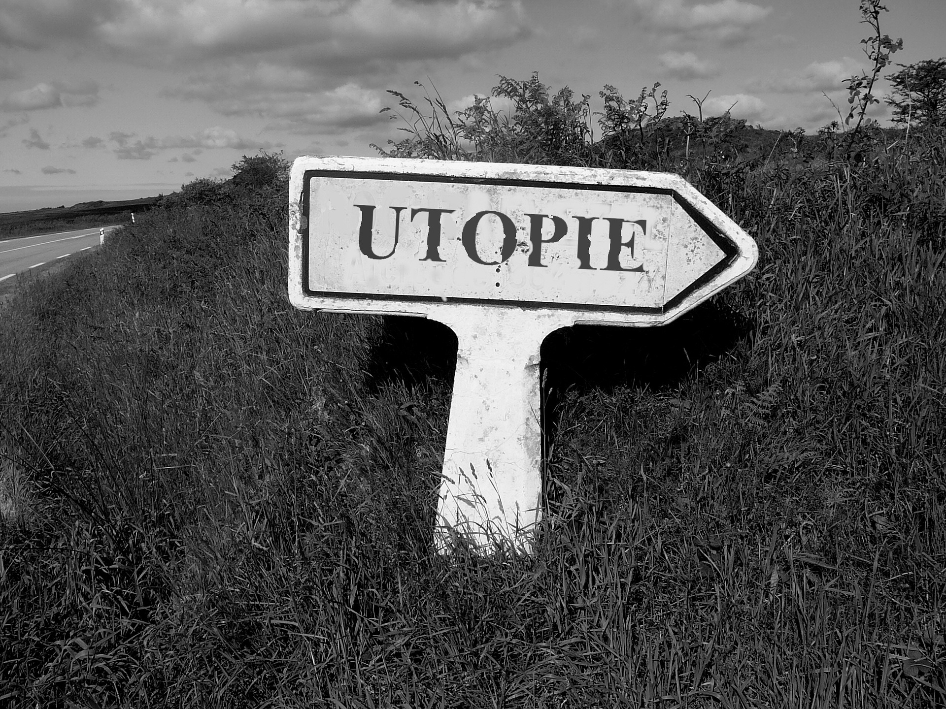 Ein Schwarz-Weiß-Bild einer Landschaft mit einer Straßenböschung, in dessen Zentrum ein Wegweiser mit der großen Aufschrift "Utopie" steht.