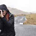 Eine warm angezogene Frau macht mit ihrer Kamera ein Foto auf einer einfachen Straße in karger Landschaft.