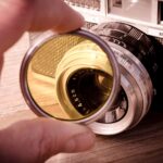 Eine Hand hält einen Gelbfilter, hinter dem eine analoge Kamera zu sehen ist.