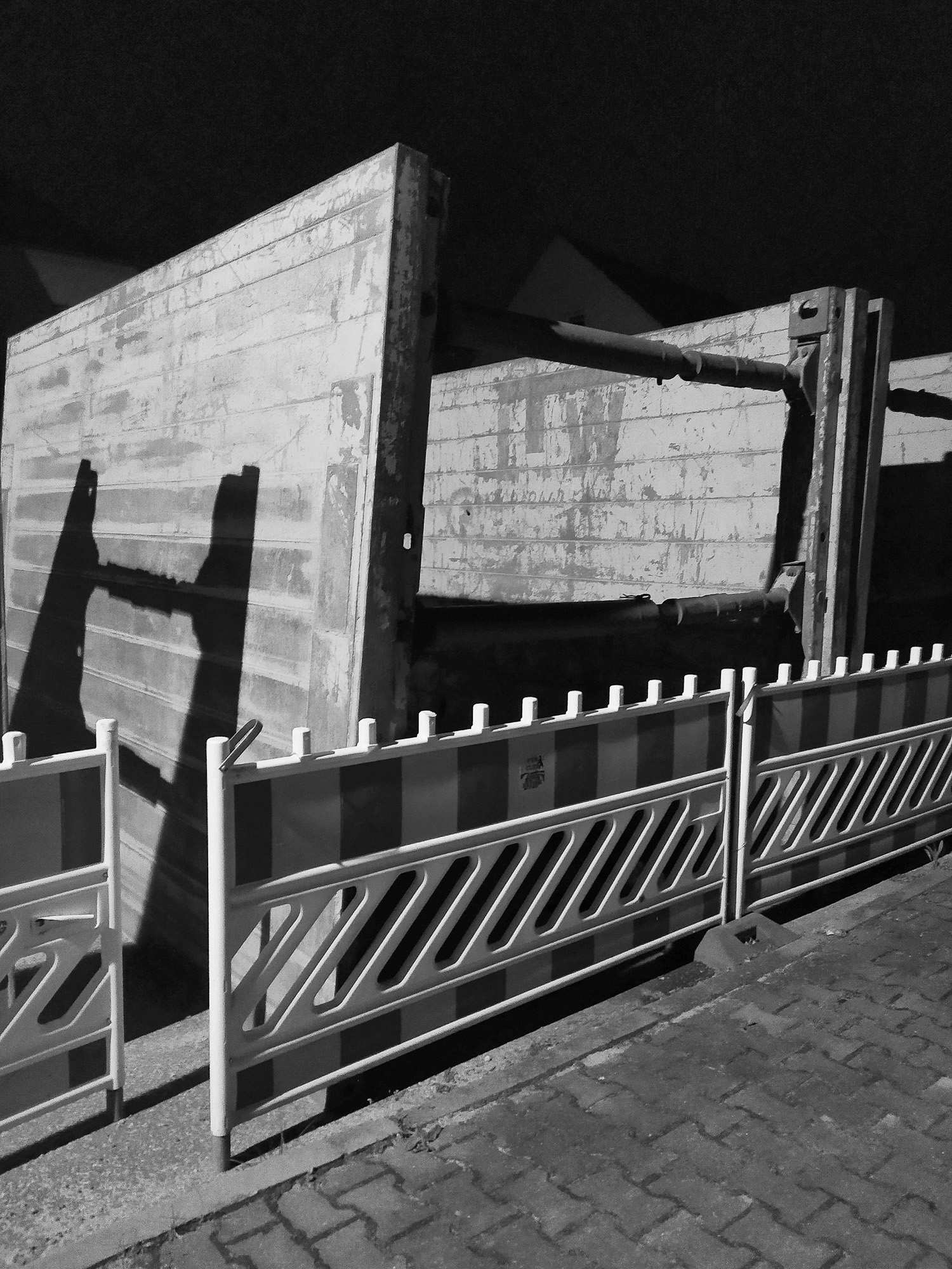 Betonbarrieren und ein provisorischer Zaun sperren nachts einen Abschnitt einer gepflasterten Straße und werfen Schatten auf den Boden. (KI generierte Beschreibung)