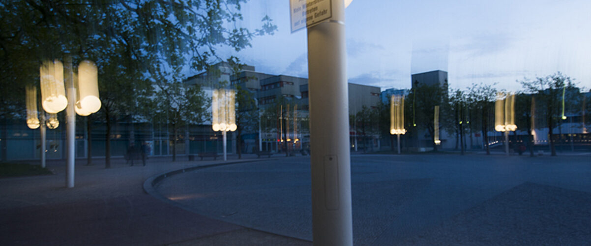 Straßenlaterne mit mehreren leuchtenden Kugeln in der Dämmerung auf einem öffentlichen Platz, die Licht auf die umliegenden Bäume und den Bürgersteig wirft.