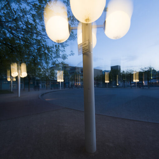 Straßenlaterne mit mehreren leuchtenden Kugeln in der Dämmerung auf einem öffentlichen Platz, die Licht auf die umliegenden Bäume und den Bürgersteig wirft.