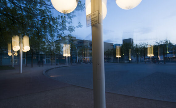 Straßenlaterne mit mehreren leuchtenden Kugeln in der Dämmerung auf einem öffentlichen Platz, die Licht auf die umliegenden Bäume und den Bürgersteig wirft.