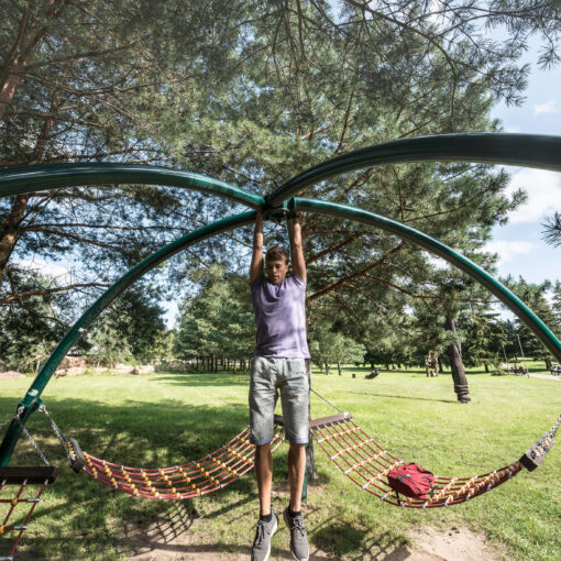 Ein junger Mann hängt an einer gebogenen Metallstange in einem Park mit Bäumen und Hängematten im Hintergrund.