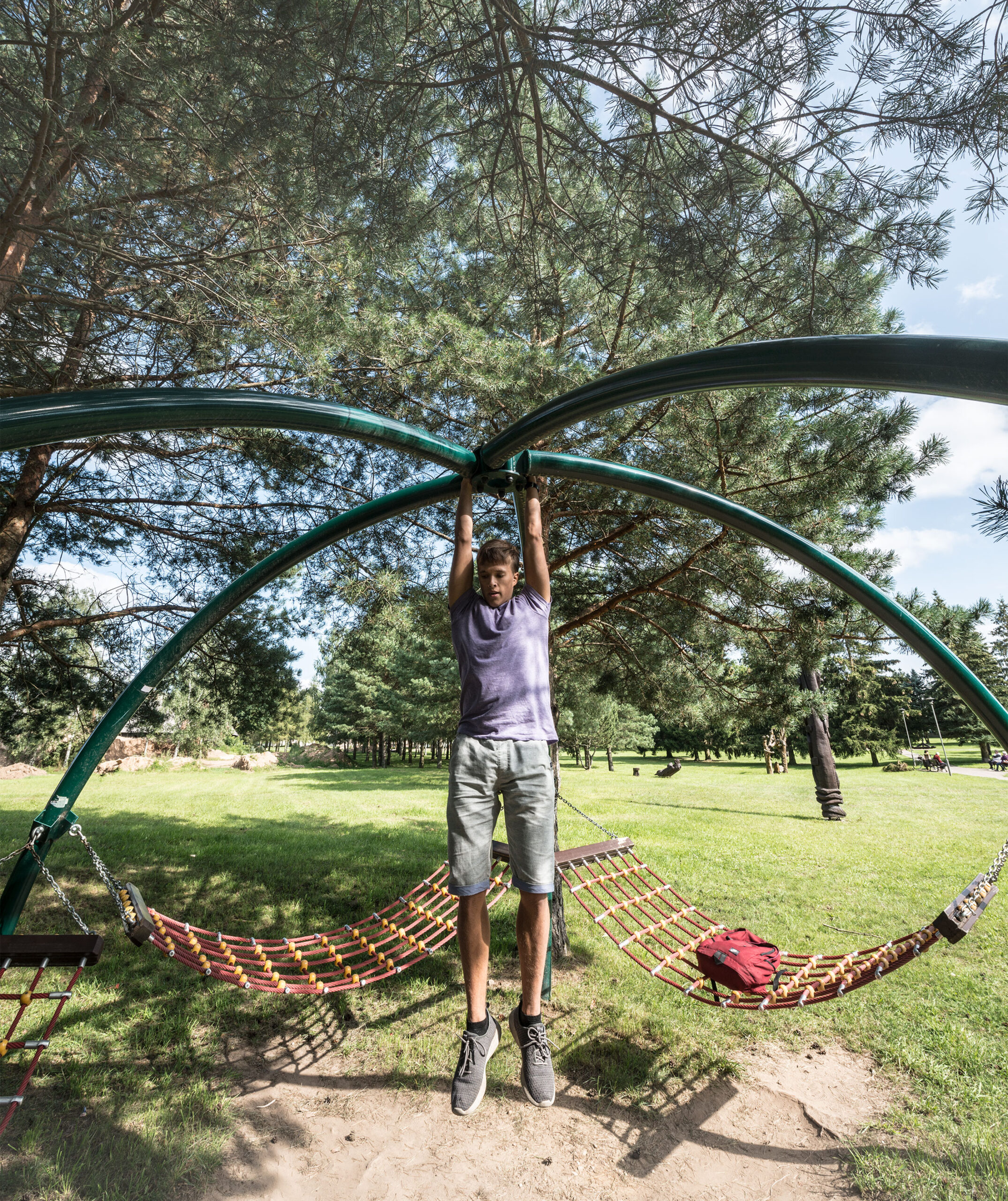 Ein junger Mann hängt an einer gebogenen Metallstange in einem Park mit Bäumen und Hängematten im Hintergrund.