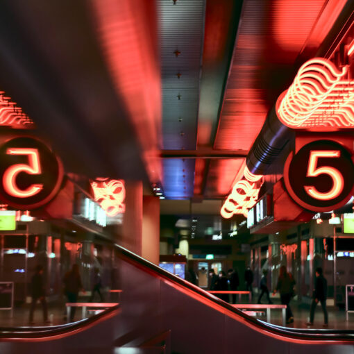 Verschwommene Ansicht eines modernen Bahnhofs mit leuchtend roten Zahlen und sich bewegenden Menschen, was eine geschäftige, dynamische Atmosphäre widerspiegelt.