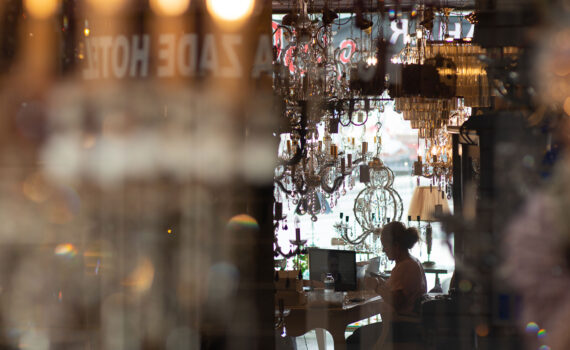 Eine Person, die in einem Café an einem Laptop arbeitet. Der Blick erfolgt durch ein Fenster mit Reflexionen von Hängelampen und unscharfen Vordergrundelementen.