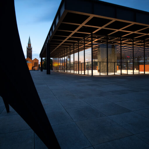 Moderne Architektur mit großen Glasflächen und eckigen Strukturen in der Abenddämmerung, im Vordergrund eine Skulptur, im Hintergrund ist eine alte Kirche zu sehen.