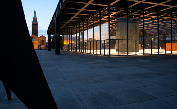 Moderne Architektur mit großen Glasflächen und eckigen Strukturen in der Abenddämmerung, im Vordergrund eine Skulptur, im Hintergrund ist eine alte Kirche zu sehen.