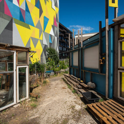 Stadtgarten mit provisorischen Gewächshäusern neben einem Gebäude mit einem farbenfrohen geometrischen Wandgemälde unter einem klaren blauen Himmel.