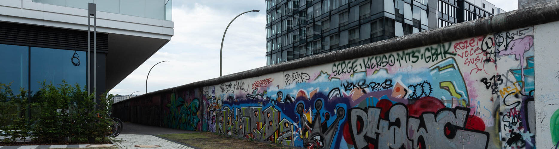 Moderne Stadtlandschaft mit einer mit Graffiti bedeckten Wand, einem gepflasterten Gehweg, zeitgenössischen Gebäuden und bewölktem Himmel.