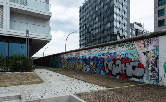 Moderne Stadtlandschaft mit einer mit Graffiti bedeckten Wand, einem gepflasterten Gehweg, zeitgenössischen Gebäuden und bewölktem Himmel.