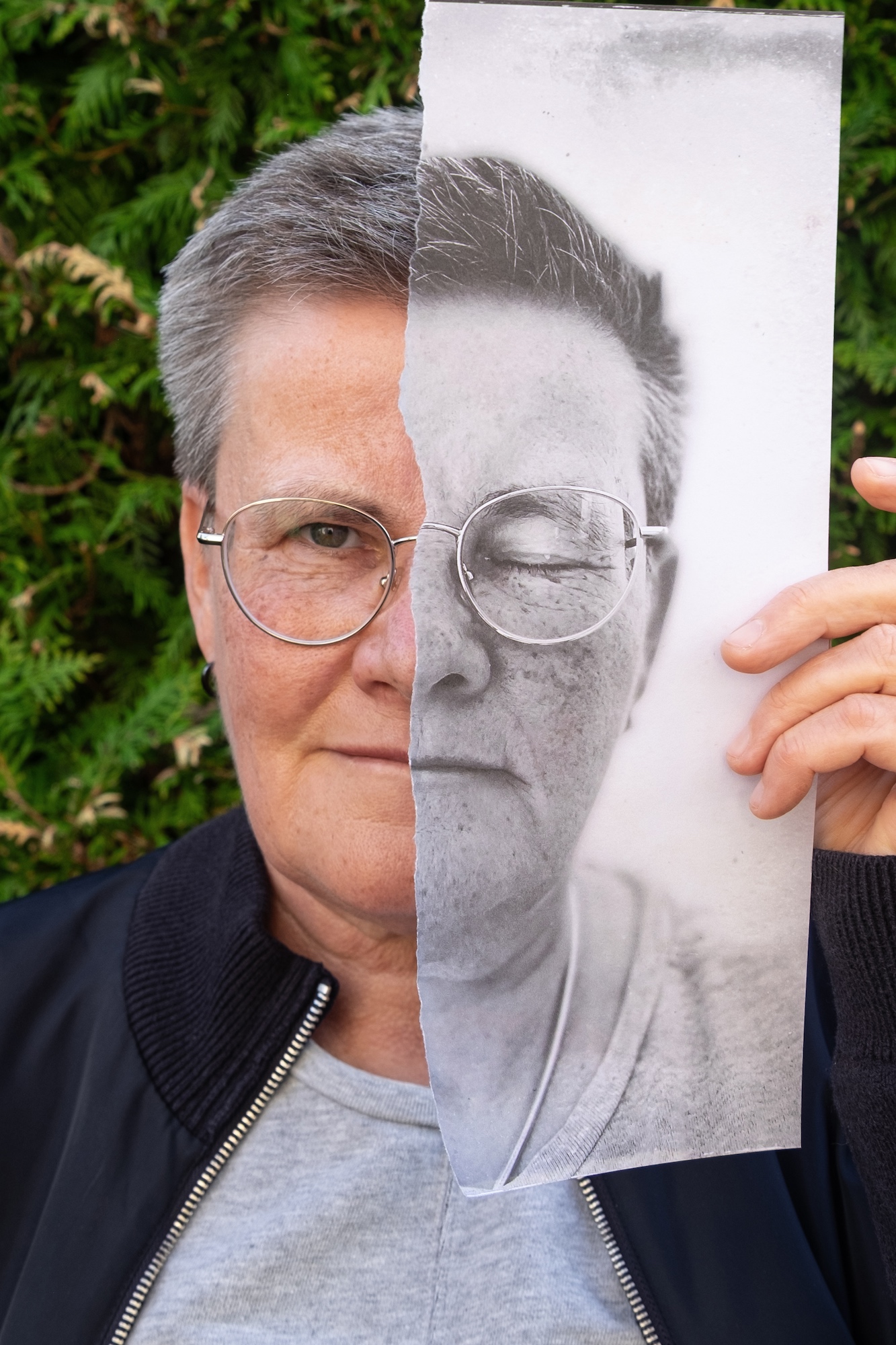 Eine ältere Person mit kurzen Haaren und Brille hält ein zerrissenes Schwarz-Weiß-Foto ihres Gesichts vor einem Hintergrund aus grünem Laub in die Höhe und richtet es an ihrem eigenen aus. (KI generierte Beschreibung)