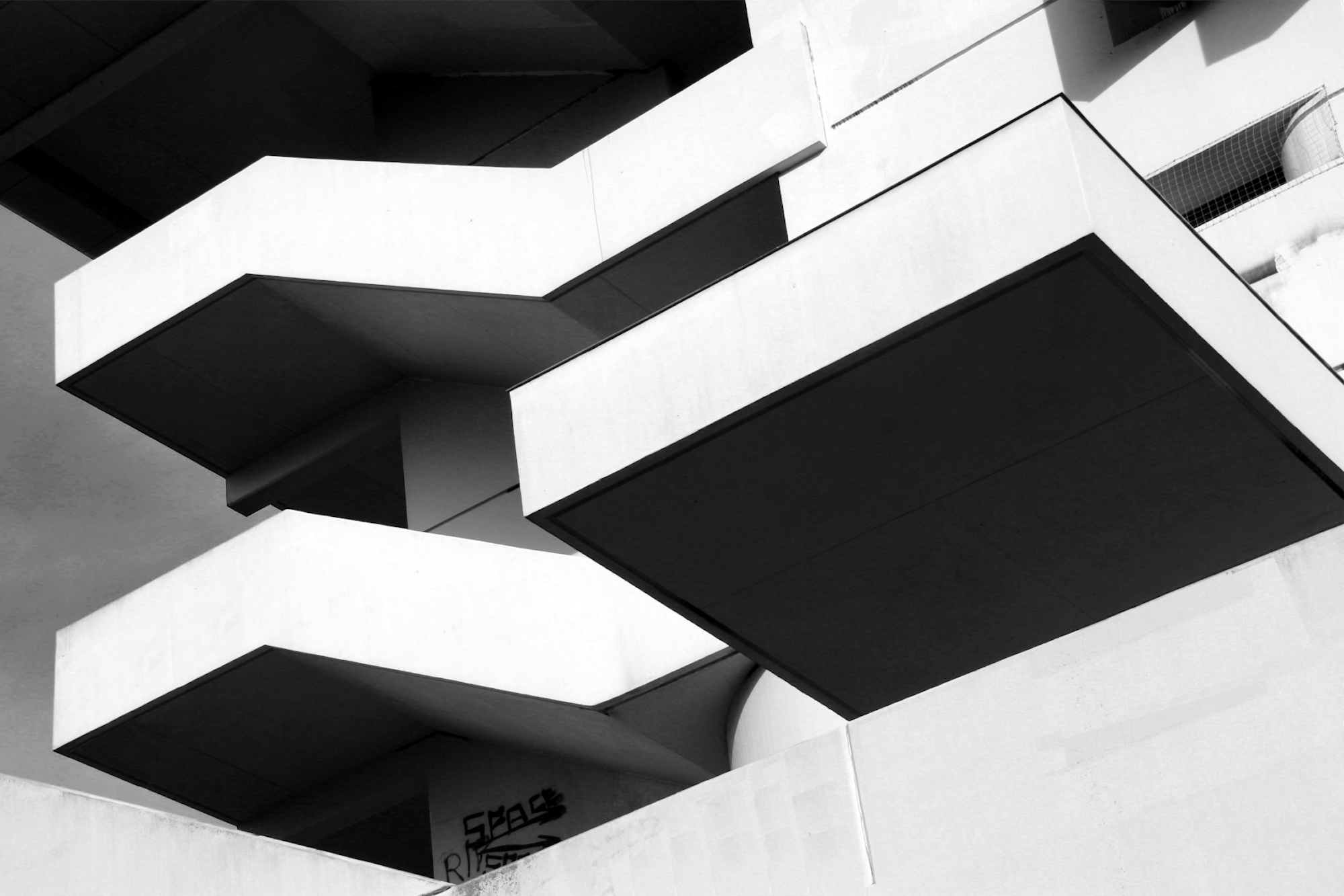 Schwarz-Weiß-Foto der rechteckigen Balkone eines modernen Gebäudes, von unten gesehen, mit scharfen Winkeln und starken geometrischen Linien. (KI generierte Beschreibung)