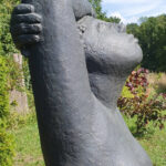 Statue im Freien, die eine Person mit erhobenem Arm und nach hinten geneigtem Kopf darstellt, in einem Garten mit grünen Bäumen und Sträuchern im Hintergrund. (KI generierte Beschreibung)