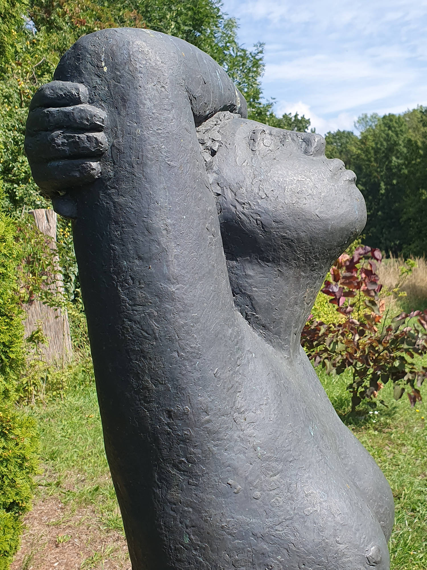 Statue im Freien, die eine Person mit erhobenem Arm und nach hinten geneigtem Kopf darstellt, in einem Garten mit grünen Bäumen und Sträuchern im Hintergrund. (KI generierte Beschreibung)