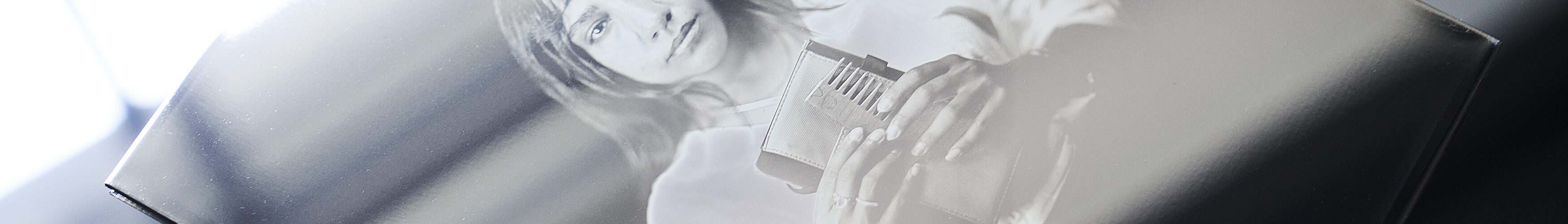 Eine Hand hält eine Glasplatte mit dem schwarz-weißen fotografischen Porträt einer Person, die einen Gegenstand in der Hand hält, wobei die Spiegelungen auf der Glasoberfläche sichtbar sind. (KI generierte Beschreibung)
