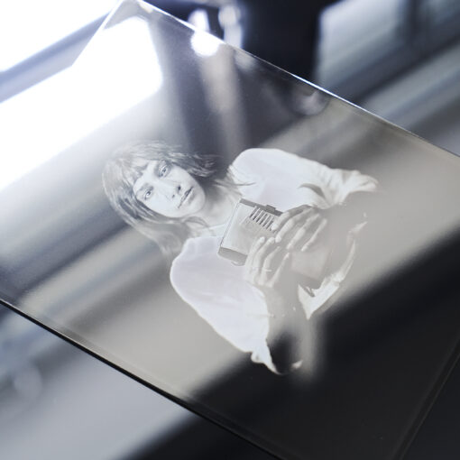 Eine Hand hält eine Glasplatte mit dem schwarz-weißen fotografischen Porträt einer Person, die einen Gegenstand in der Hand hält, wobei die Spiegelungen auf der Glasoberfläche sichtbar sind. (KI generierte Beschreibung)