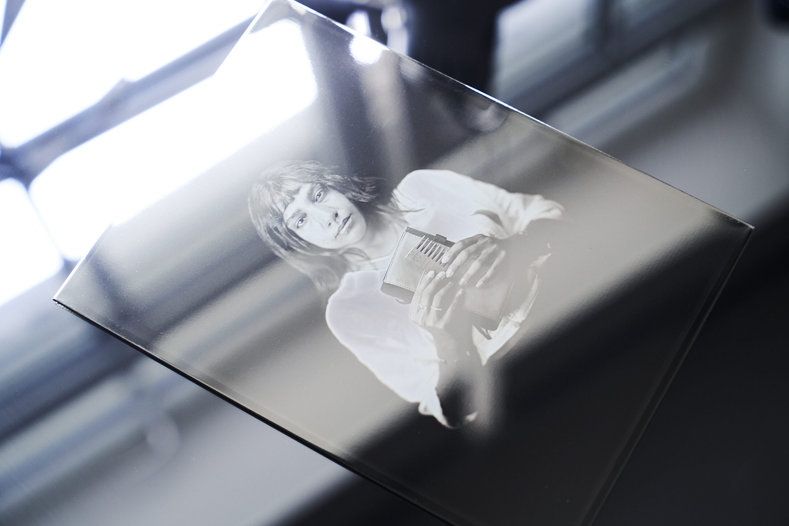 Eine Hand hält eine Glasplatte mit dem schwarz-weißen fotografischen Porträt einer Person, die einen Gegenstand in der Hand hält, wobei die Spiegelungen auf der Glasoberfläche sichtbar sind. (KI generierte Beschreibung)