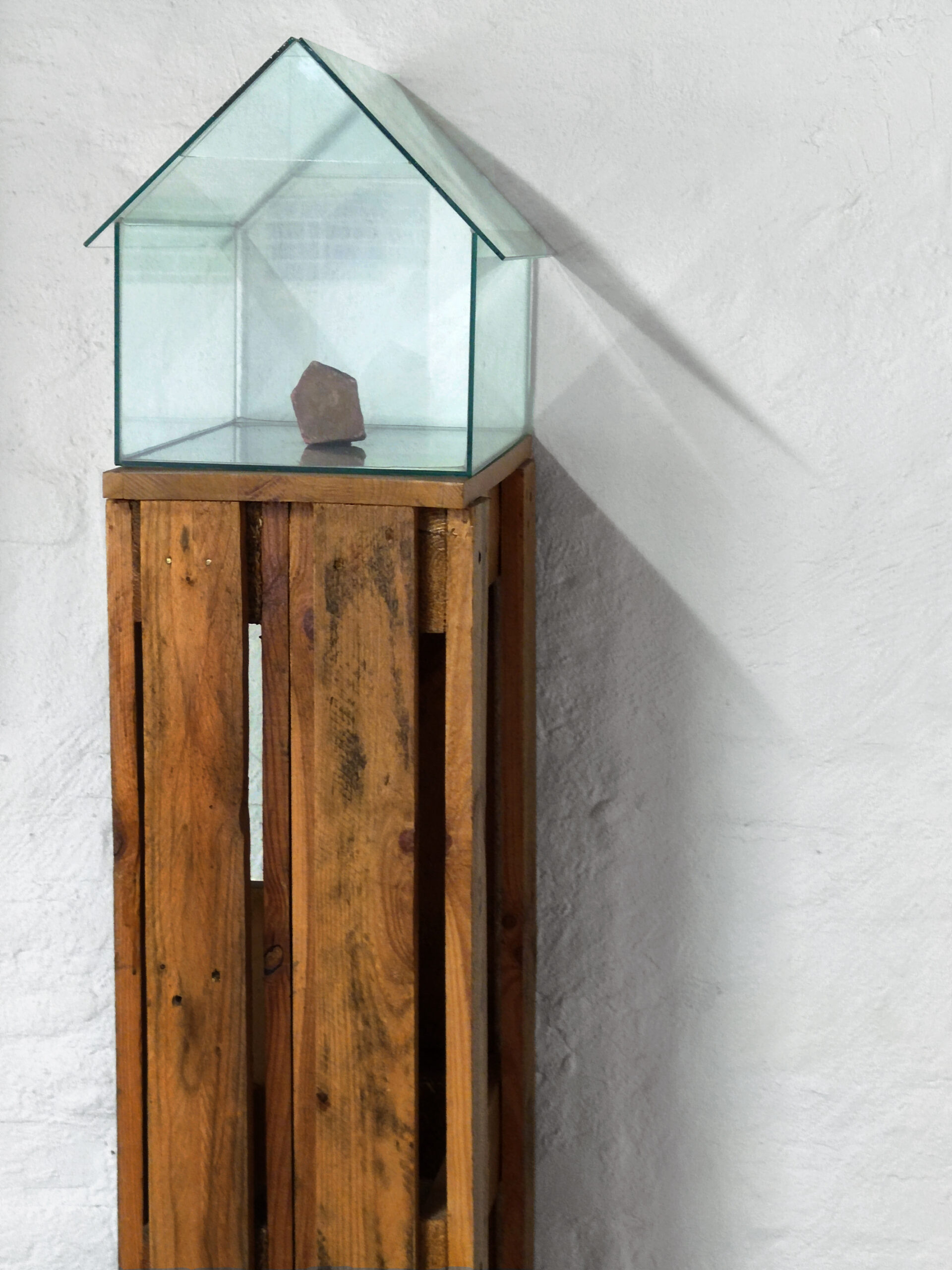 Ein kleiner Stein wird in einer Glasvitrine in Form eines Hauses ausgestellt, die auf einem hohen Holzsockel vor einer weißen Wand steht. (KI generierte Beschreibung)