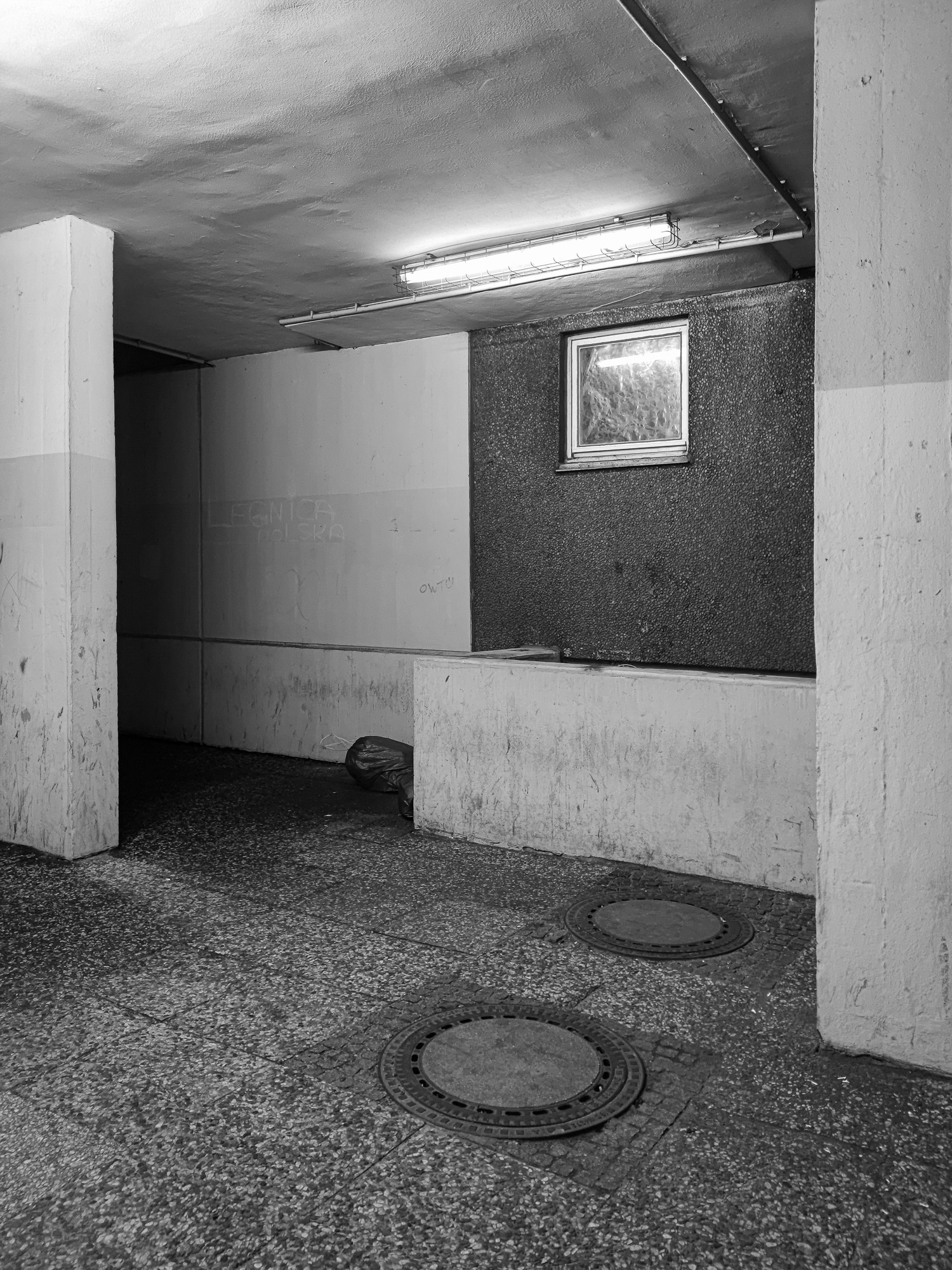 Ein schwach beleuchteter, leerer Flur mit Betonwänden und -boden. In einer Wand befindet sich ein kleines Fenster mit einer Leuchtstoffröhre darüber. Auf dem Boden liegen zwei runde Metallgitter. (KI generierte Beschreibung)