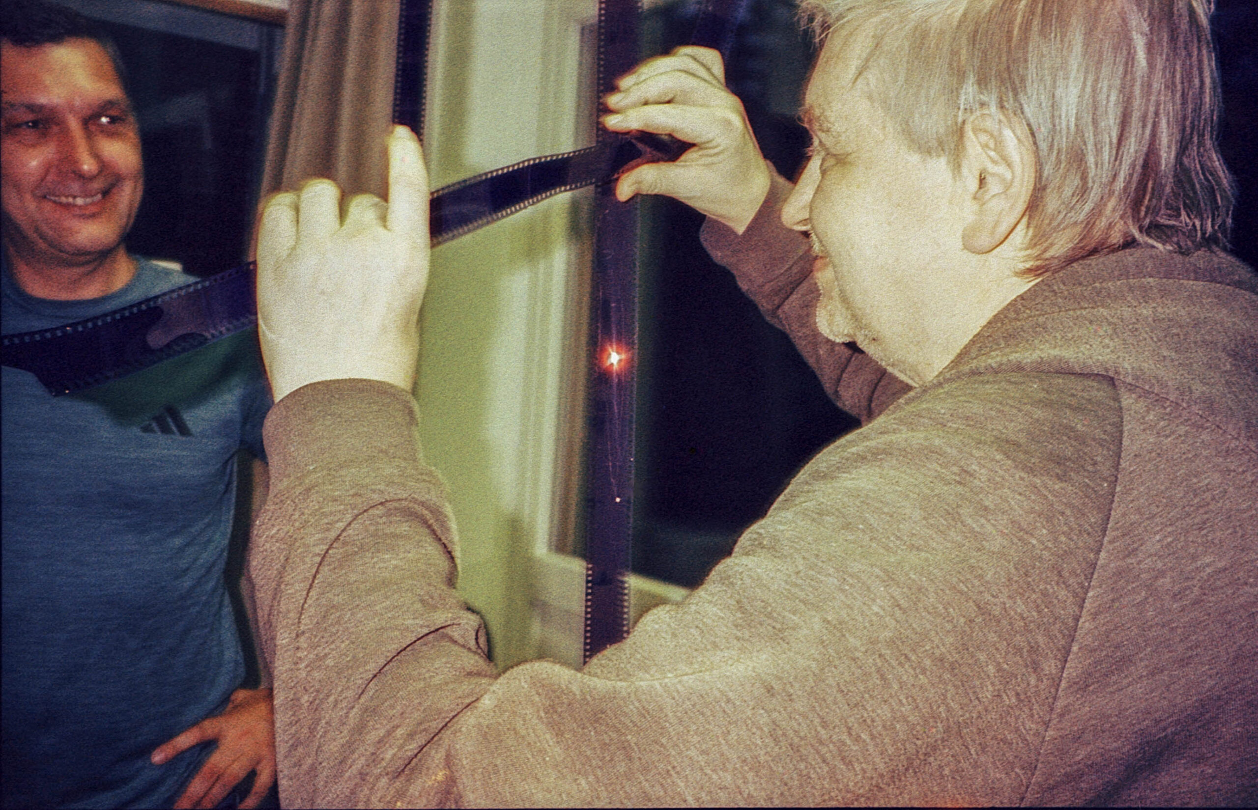 Zwei Personen untersuchen in einem Innenraum einen Fotostreifen; eine hält den Film gegen das Licht, während die andere lächelnd zusieht. (KI generierte Beschreibung)
