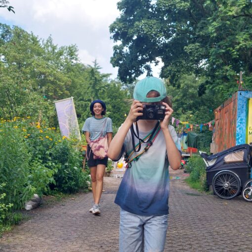 Ein Kind mit einer Mütze und einer Kamera in der Hand steht auf einem Weg, während ein Erwachsener hinter ihm geht. Fahrräder, ein Kinderwagen und Grünflächen umgeben den Bereich. (KI generierte Beschreibung)