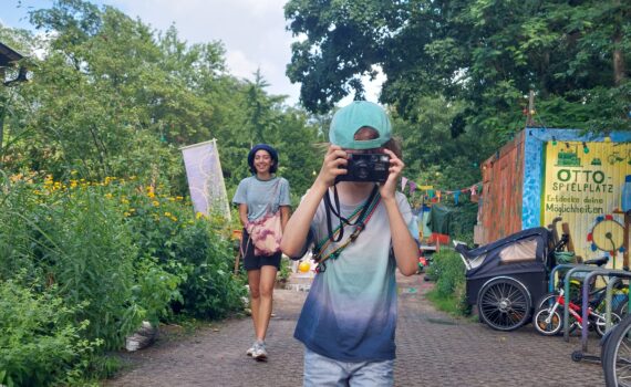 Ein Kind mit einer Mütze und einer Kamera in der Hand steht auf einem Weg, während ein Erwachsener hinter ihm geht. Fahrräder, ein Kinderwagen und Grünflächen umgeben den Bereich. (KI generierte Beschreibung)