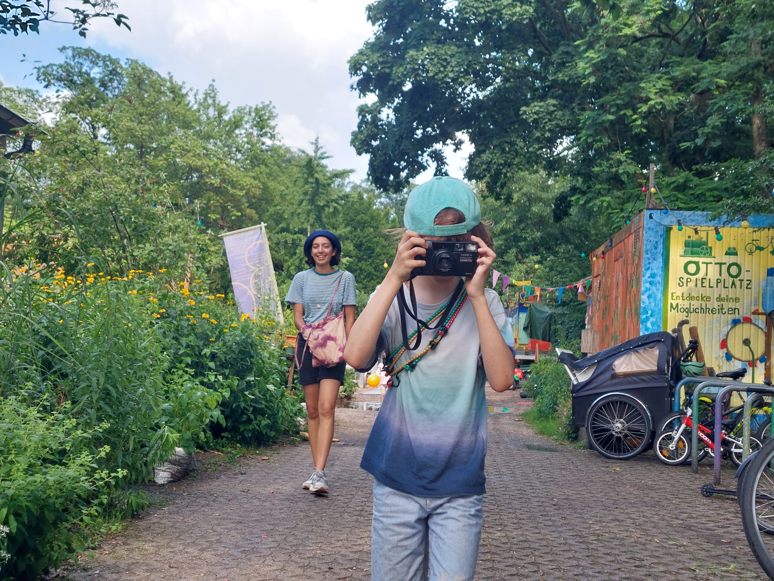 Ein Kind mit einer Mütze und einer Kamera in der Hand steht auf einem Weg, während ein Erwachsener hinter ihm geht. Fahrräder, ein Kinderwagen und Grünflächen umgeben den Bereich. (KI generierte Beschreibung)