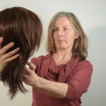 Eine Frau hält und betrachtet eine braune Perücke vor einem schlichten Hintergrund. (KI generierte Beschreibung)