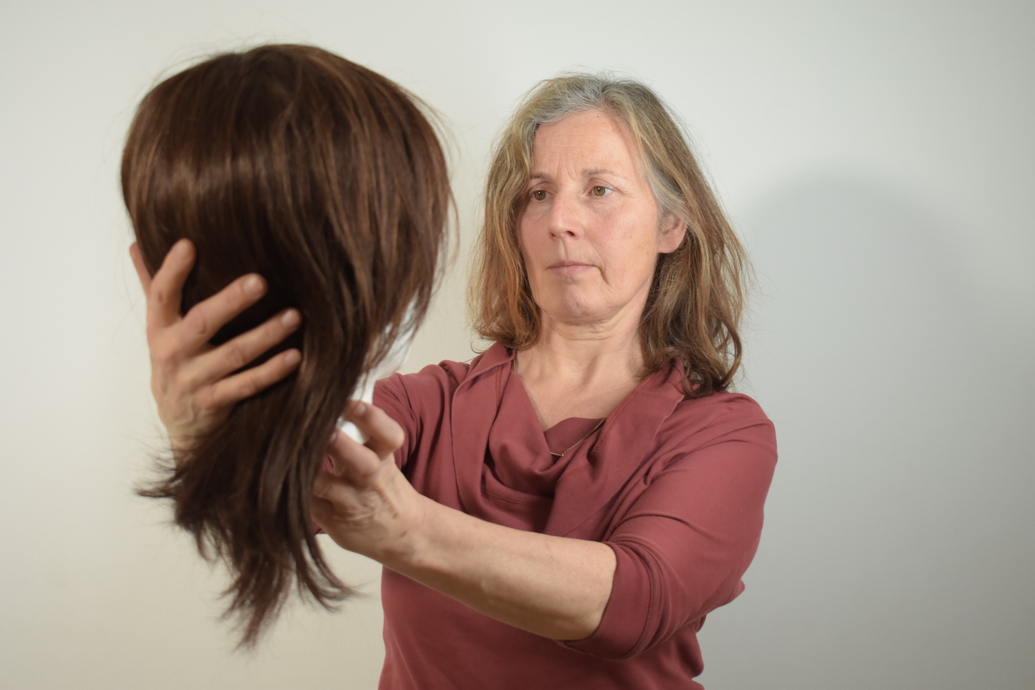 Eine Frau hält und betrachtet eine braune Perücke vor einem schlichten Hintergrund. (KI generierte Beschreibung)