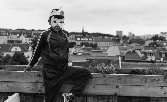 Ein Junge mit einer Papierkrone und einer Maske steht auf einem Dach und blickt auf die Stadt hinunter. (KI generierte Beschreibung)